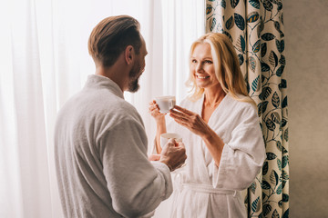 Obraz na płótnie Canvas beautiful happy mature couple in bathrobes drinking coffee and smiling each other in hotel room