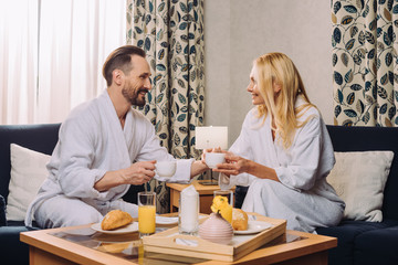 happy middle aged couple in bathrobes drinking coffee during breakfast in hotel room