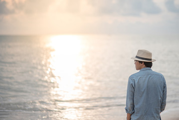 Obraz premium Young Asian man with jean shirt and hat standing on the beach and looking beautiful sunset, vacation time and summer holiday concepts