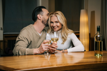 handsome mature man whispering something to smiling woman while drinking wine in hotel restaurant