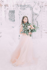 Bride in long pink dress holding wedding bouquet near the wooden house in winter mountains