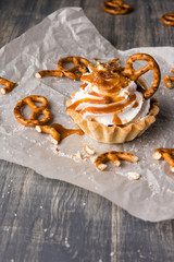 Homemade cupcake with salted caramel and pretzels. Sweet cake with syrup and cream cheese on baking paper and wooden table.