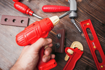 A toy drill in a man's hand and other tools on a wooden background