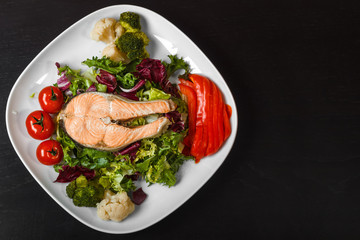 Salmon steak on a mix-salad cushion, fresh peppers and tomatoes, boiled cauliflower and broccoli on a white square plate on a dark wooden table. Top view. Copy Space