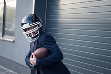 young businessman in suit and rugby helmet with ball in hands standing on street