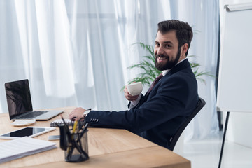 cheerful businessman in suit drinking coffee at workplace with laptop in office
