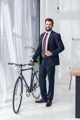 Fototapeta na wymiar smiling businessman in suit standing near bicycle in office