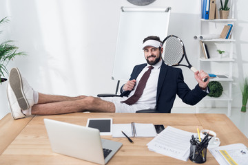 young businessman in cap with tennis racket resting at workplace in office