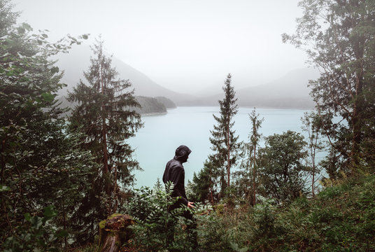 Mann mit schwarzer Jacke wandert im Regen durch die Berge mit See im Hintergrund