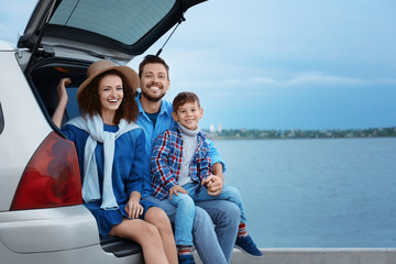 Young family with boy sitting in car trunk near river