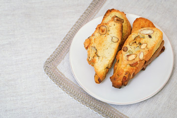 Italian dry cookies biscotti with nuts on white plate. Cantucci folded in a pile for breakfast on tablecloth of fabric. Selective focus. Copy space.