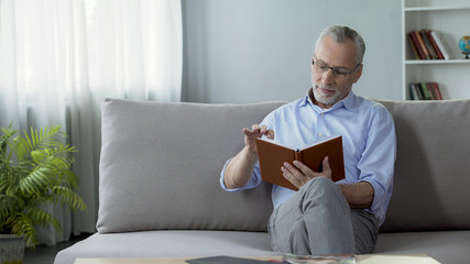 Happy old man sitting on couch and reading weekend plan, hobby and free time