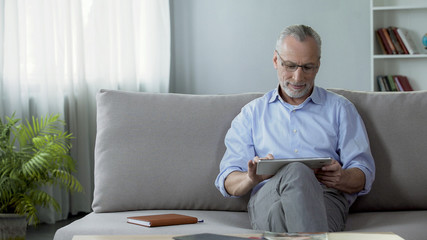 Smiling adult person sitting on couch and viewing family photos on tablet