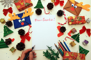 Fototapeta na wymiar merry christmas text, seasonal greetings card sign. hands holding pencil and writing a letter wish list or greeting flat lay with space for text. hand craft and presents and ornaments on white wood