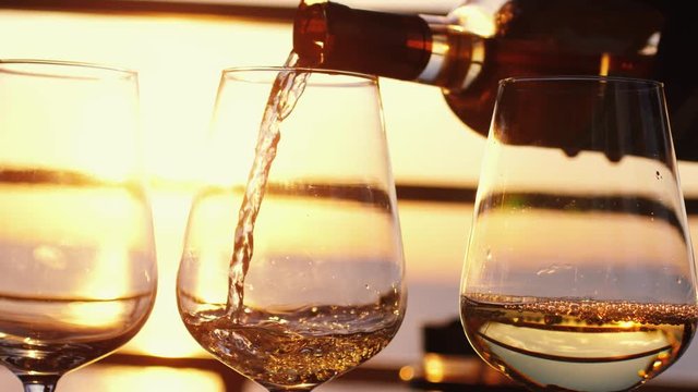 Pouring white wine in four glasses at amazing sunset by the sea in beach cafe before partying in slow motion. 3840x2160, 4k