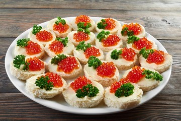 White plate on the wooden table, full of little canapees with butter,red caviar and decorated with fresh green parsley leaves. Tasty appetizer foe alcohol catering or restaurant buffet