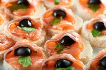 Little bruschetti with smoked salmon rolls and black olives on the white bread,covered with butter and decorated with fresh green parsley leaves.