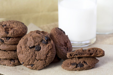 Chocolate chip cookies on brown paper and sack cloth with glass of milk and paper bag on white background.