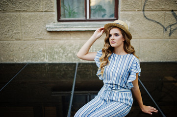 Portrait of a fabulous young woman wearing striped overall and hat sitting on black shiny surface next to the building.