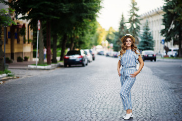 Portrait of a beautiful model in striped overall posing with hat and a backpack on a street with trees in a town.