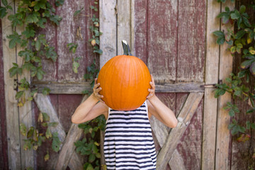 Playful girl holding pumpkin against face while standing against wooden wall at yard