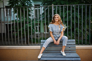 Fabulous young attractive model in striped overall sitting on a bench outdoor with trees on the background.