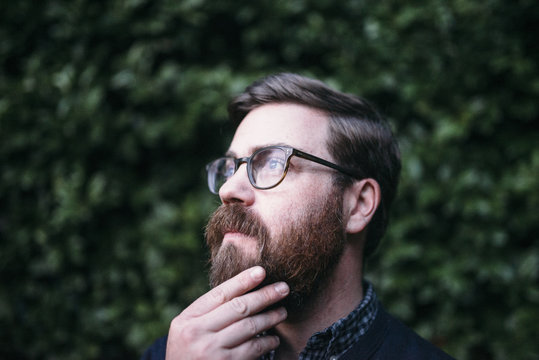 Thoughtful hipster with hand on chin looking away while wearing eyeglasses against hedge