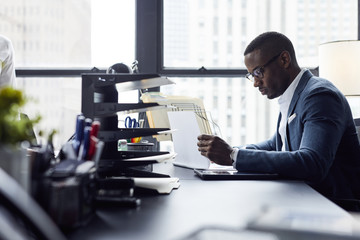 Serious businessman looking at documents while sitting against window in office