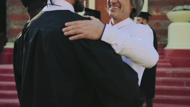 Gray-haired handsome man congratulating young man in special clothes with the getting a diploma at the graduation. Outside