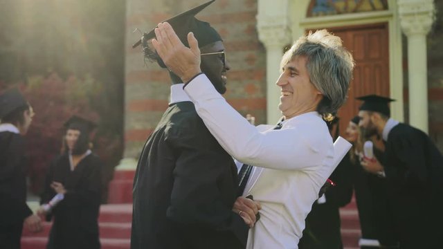 Handsome man congratulating young African-American man in special clothes with the getting a diploma at the graduation. Outdoor