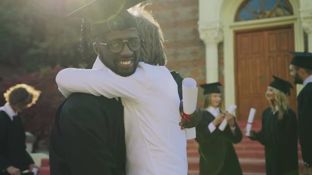 Portrait shot of the young male African American graduate in the cap getting congratulations from his proffessor near the University entrance. Outdoors