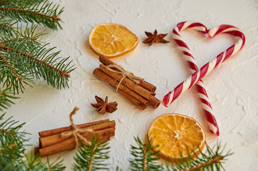 Red heart of sweet candy cones with christmas spices - cinnamon sticks, anise stars, dried oranges on the white concrete background. New year food decorations with green Christmas tree branch