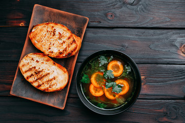 Vegetable soup decorated with sliced carrots and green parsley and toasts, flat lay with copy space on dark wooden table. Appetizing first courses menu, kitchen background, restaurant concept