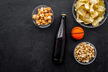 Snacks for watching sport matches and games on TV. Crisps, popcorn, rusks near drink and ball on black background top view copyspace