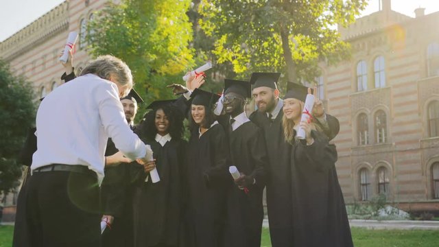 Back view on the gray-haired man taking a photo portrait on the black smartphone of the multi ethnical graduates in traditional black clothes and caps with diplomas in hands. Outdoors