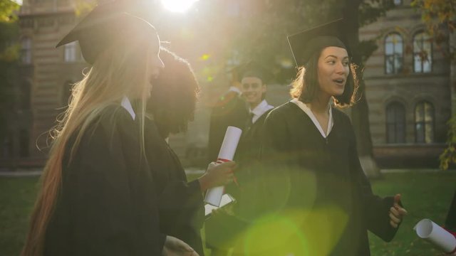 Pretty multi ethnical graduated girls in gowns and caps holding their diplomas and talking joyfully near the University. Guys on the background. Outdoors