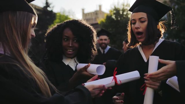 Charming young female graduates in traditional gowns and caps unwrapping diploma and saying WOW when looking at it. Guys on the background. Outside