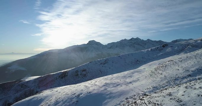 Forward aerial top view over winter snowy mountain with ski tracks in sunny day with clouds.Backlight white Alps mountains snow season establisher with sun shine.4k drone flight establishing shot