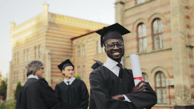 Portrait of the African American happy male graduate posing to the camera and smiling sincerely in front of the University. Graduates with professor on the background. Outside