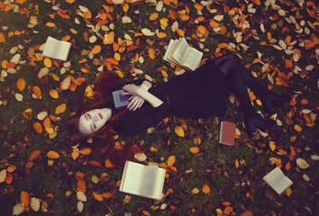 Beautiful red-haired girl with books lies on the grass in an autumn forest, top view. Autumn...