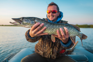 Happy young angler holds the pike fish (Esox lucius) sitting in a boat