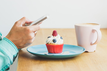 Cupcake with raspberry and blueberry on the plate, cup of cappuccino and woman's hand with a phone