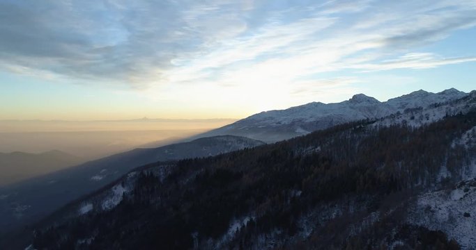 Backward aerial top view over winter snowy mountain and woods forest at sunset or sunrise.Blue hour dusk or dawn twilight Alps mountains snow season establisher.4k drone flight establishing shot
