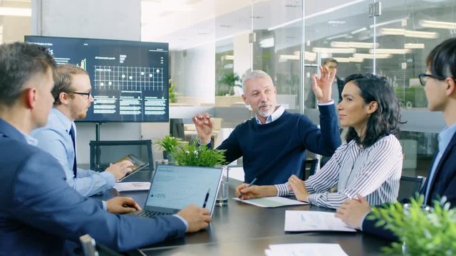 Senior Executive Explains Company's Vision and Potential to His Employees. They are Sitting at Big Table in Meeting Room. TV Screen on the Wall Shows Corporate Growth. RED EPIC-W 8K Camera.