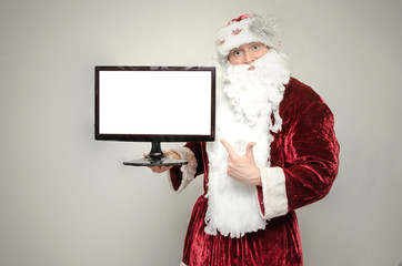 Santa claus man holds in hands a computer monitor with blank screen and showing on it with his index finger isolated on gray background.
