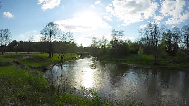 Timelapse nature. Counter flow of a small river. The water reflects the clouds and the bright sun, which through the gaps appears from behind the clouds. A calm spring landscape with a young greenery.