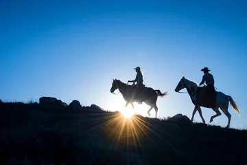 Foto auf Acrylglas Silhouetted Western Cowboy and Cowgirl on horseback against a blue sky with sun flare at horizon © Frank