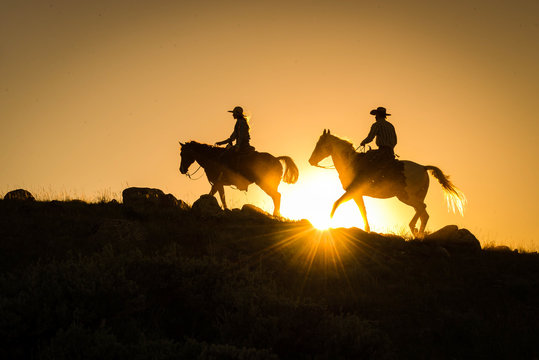 Silhouetted Western Cowboy And Cowgirl On Horseback Against Yellow Sunset 