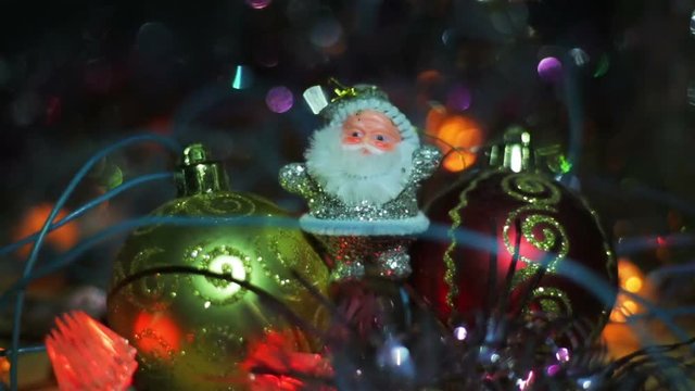 Miniature Santa Claus figure between two toy Hanging Baubles for a Christmas tree. Silver Santa on a wooden surface surrounded by flashing lights.
