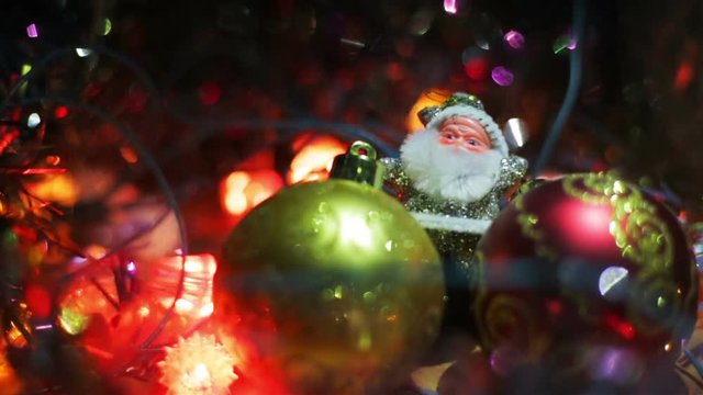Miniature Santa Claus figure between two toy Hanging Baubles for a Christmas tree. Silver Santa on a wooden surface surrounded by flashing lights.